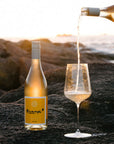 Bottle of Orange* in Colour Margaret River Organic wine being poured into a glass on a rocky surface with a scenic background.