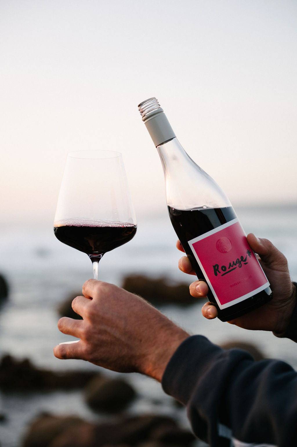 Person holding a bottle and glass of Blind Corner Organic Margaret River Rouge wine with a scenic coastal background