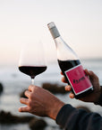 Person holding a bottle and glass of Blind Corner Organic Margaret River Rouge wine with a scenic coastal background