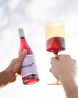 Two hands holding a bottle of Organic Nouveau and a glass of red wine against a light background