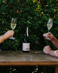 Two people clinking glasses of Blind Corner Organic and Biodynamic Margaret River Cremant on a wooden table outdoors.