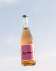 A bottle of Blind Corner Organic and Biodynamic Margaret River Petillant with a pink label, slightly hazy pink liquid, and a visible gas bubble, against a blue sky background.