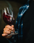 Person holding a glass of Blind Corner Organic and Biodynamic Margaret River Rouge wine with a blurred background of barrels