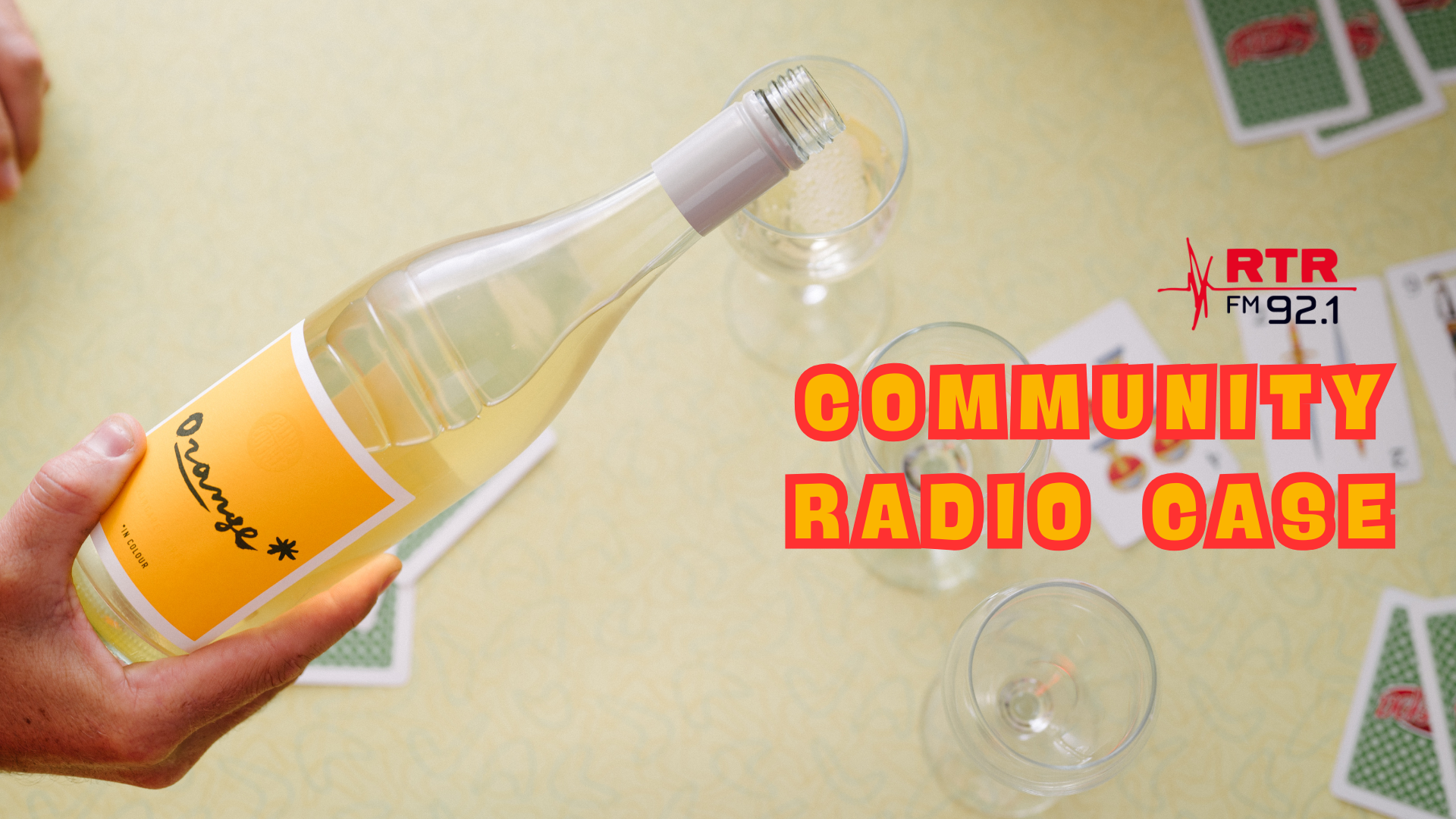 Bottle of Orange* in Colour wine held by a hand with playing cards and glasses on a yellow background, featuring RTR FM21 branding.