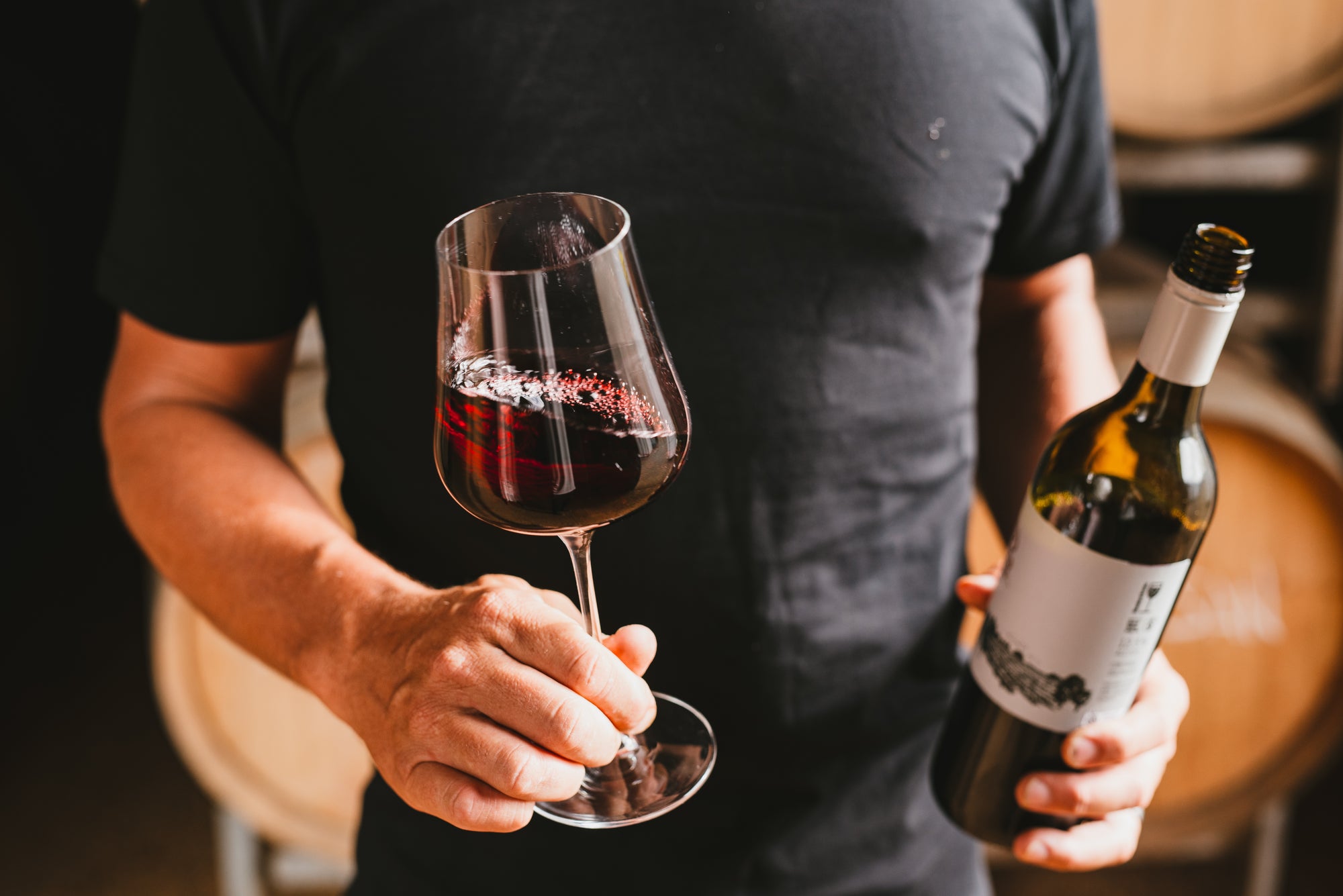 Person holding a glass of organic red wine and a bottle in a dimly lit setting