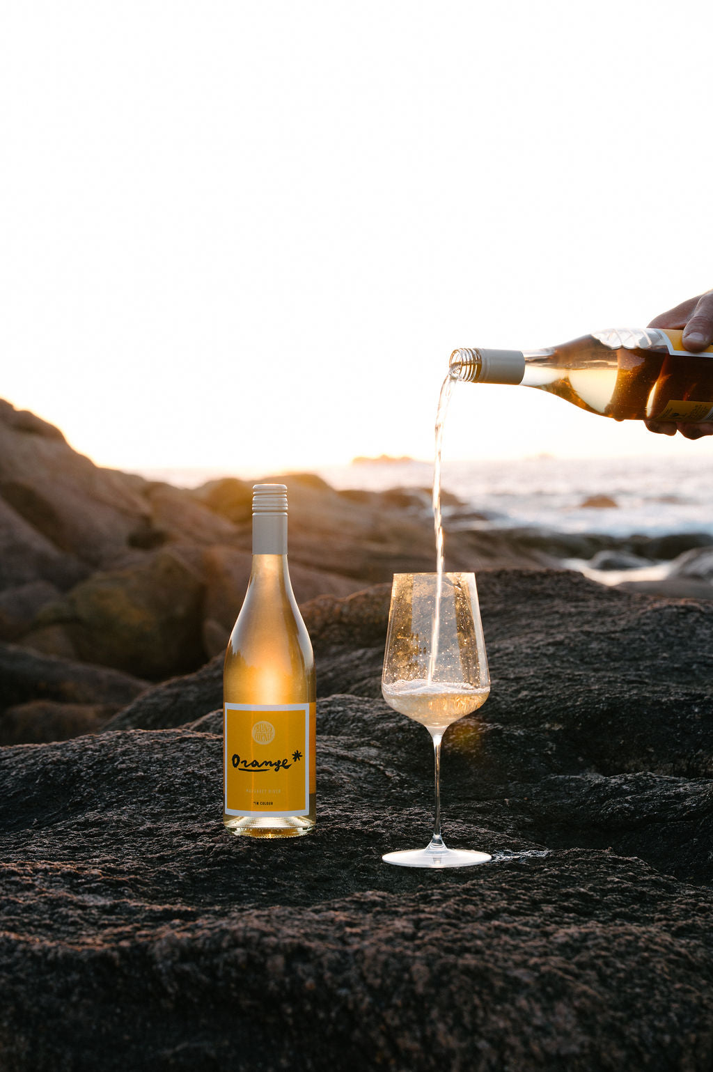 Bottle of Orange* in Colour Margaret River Organic wine being poured into a glass on a rocky surface with a scenic background. 