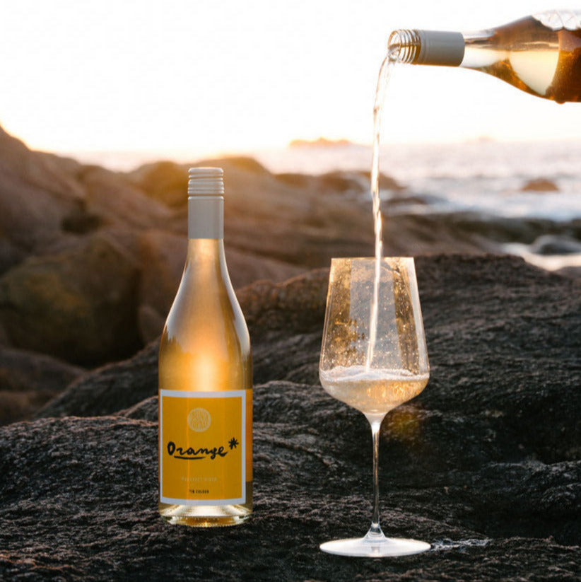 Bottle of Orange* in Colour Margaret River Organic wine being poured into a glass on a rocky surface with a scenic background. 