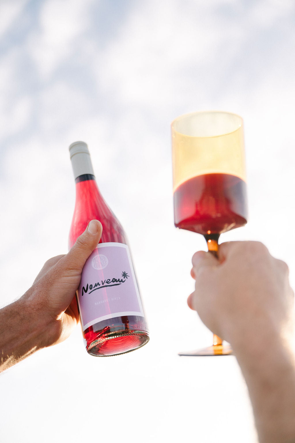 Two hands holding a bottle of Organic Nouveau and a glass of red wine against a light background