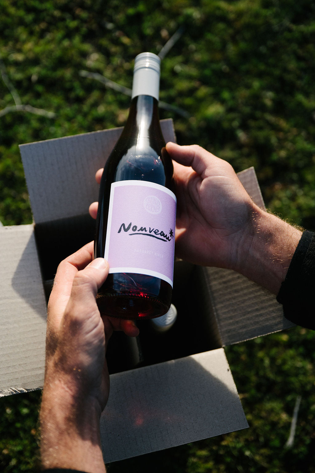 Person holding a bottle of Blind Corner Organic Nouveau red wine with a purple label outdoors. The Wine Club 4-pack box is in the background