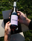 Person holding a bottle of Blind Corner Organic Nouveau red wine with a purple label outdoors. The Wine Club 4-pack box is in the background