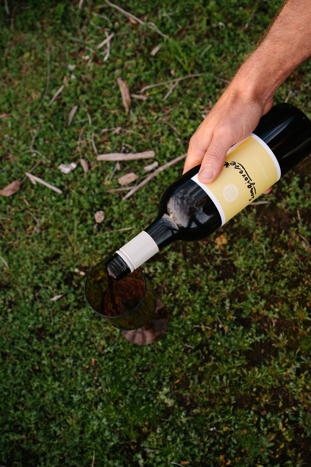 Hand pouring a bottle of Blind Corner Organic wine with a yellow label into a vintage glass on a grassy background