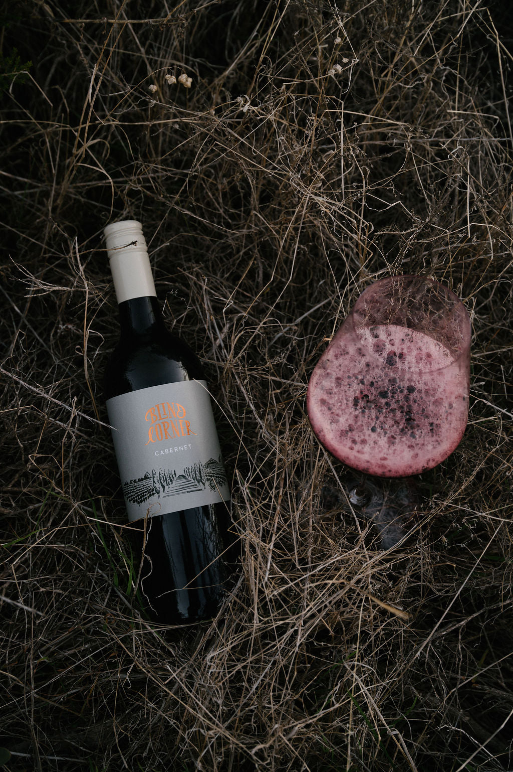 Blind Corner Organic Margaret River Cabernet bottle with a label on a dark, dried-grass surface next to a freshly poured glass of wine