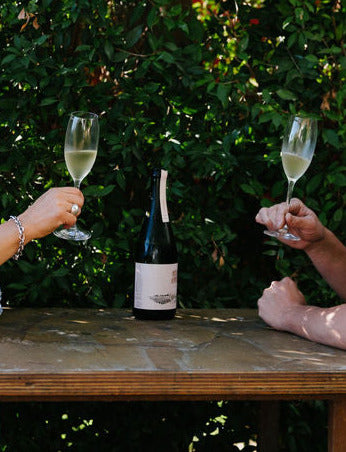 Two people clinking glasses of Blind Corner Organic and Biodynamic Margaret River Cremant on a wooden table outdoors.