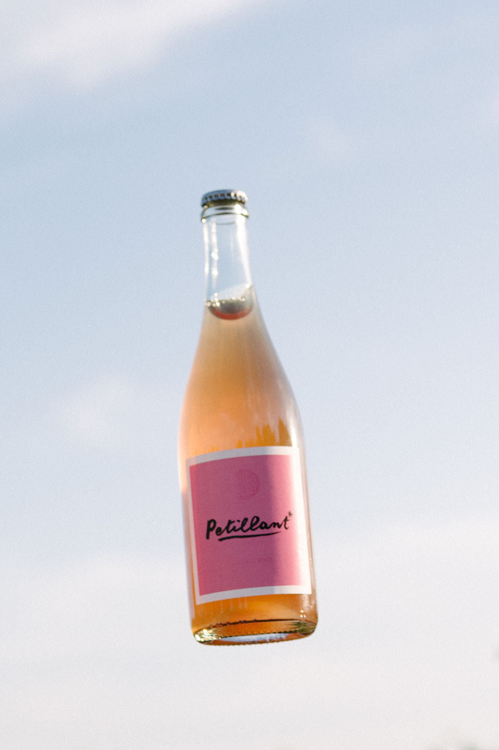 A bottle of Blind Corner Organic and Biodynamic Margaret River Petillant with a pink label, slightly hazy pink liquid, and a visible gas bubble, against a blue sky background.