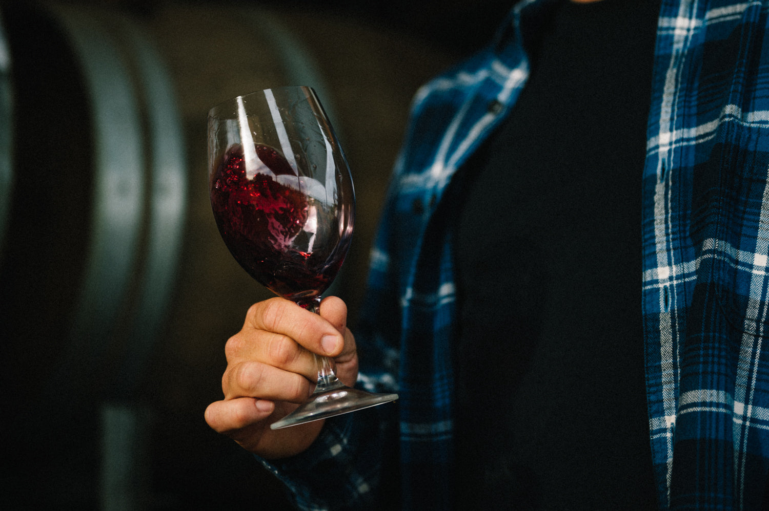 Person holding a glass of Blind Corner Organic and Biodynamic Margaret River Rouge wine with a blurred background of barrels