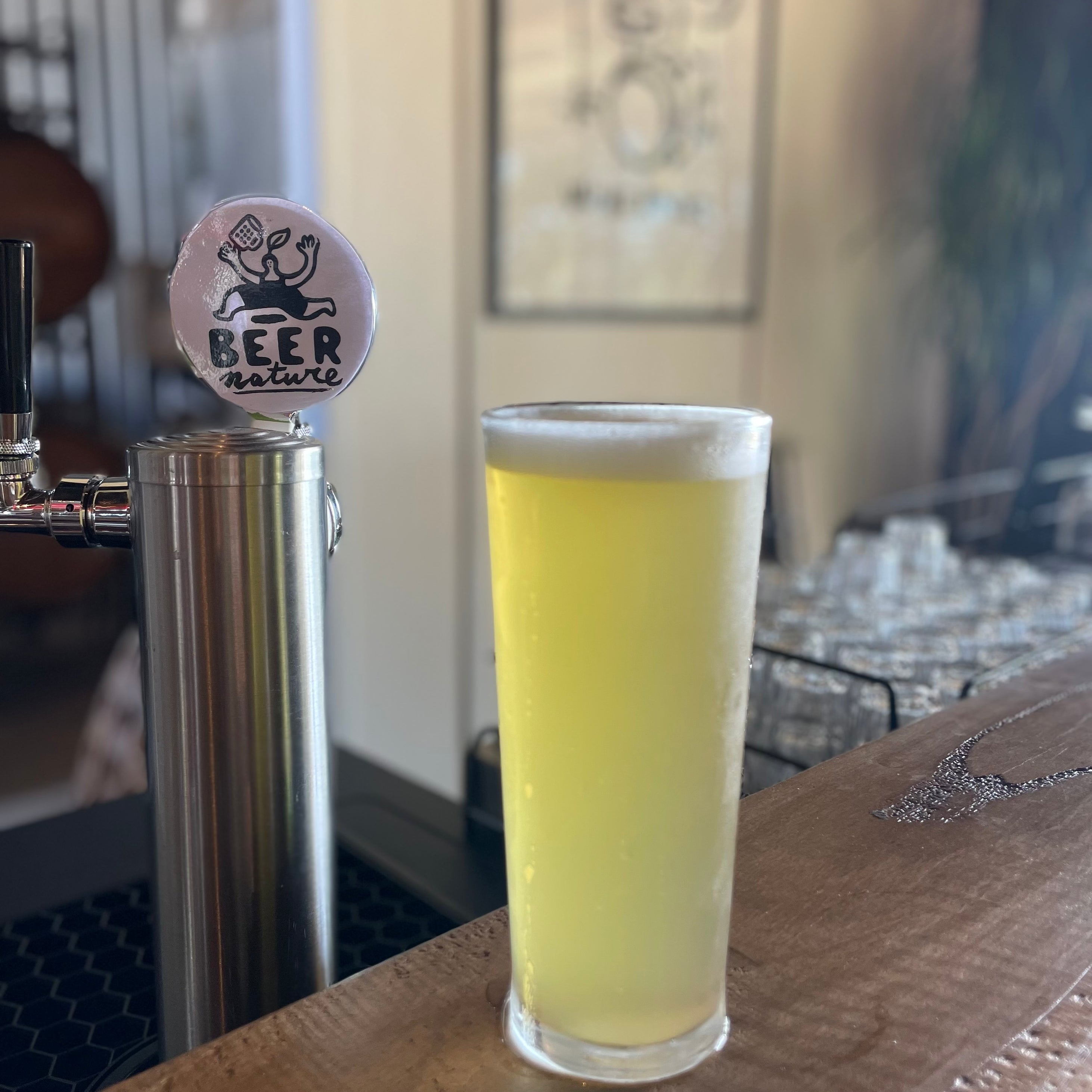 Glass of beer with a 'Beer Nature' tap handle on a bar counter.