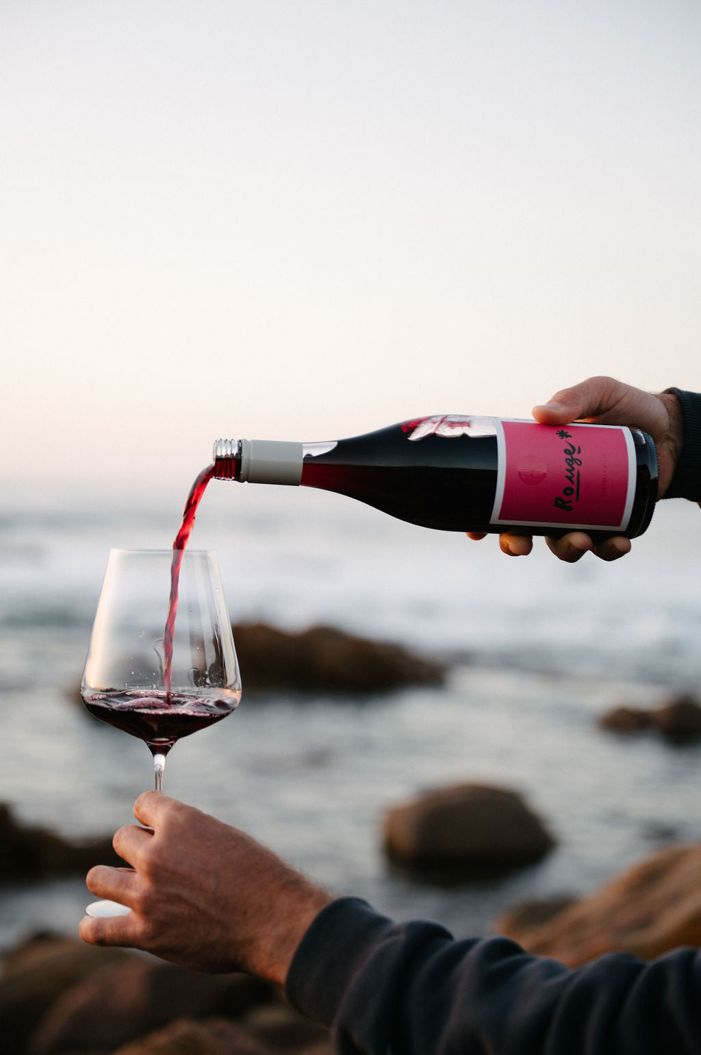 Person pouring red wine from a bottle into a glass by the ocean.