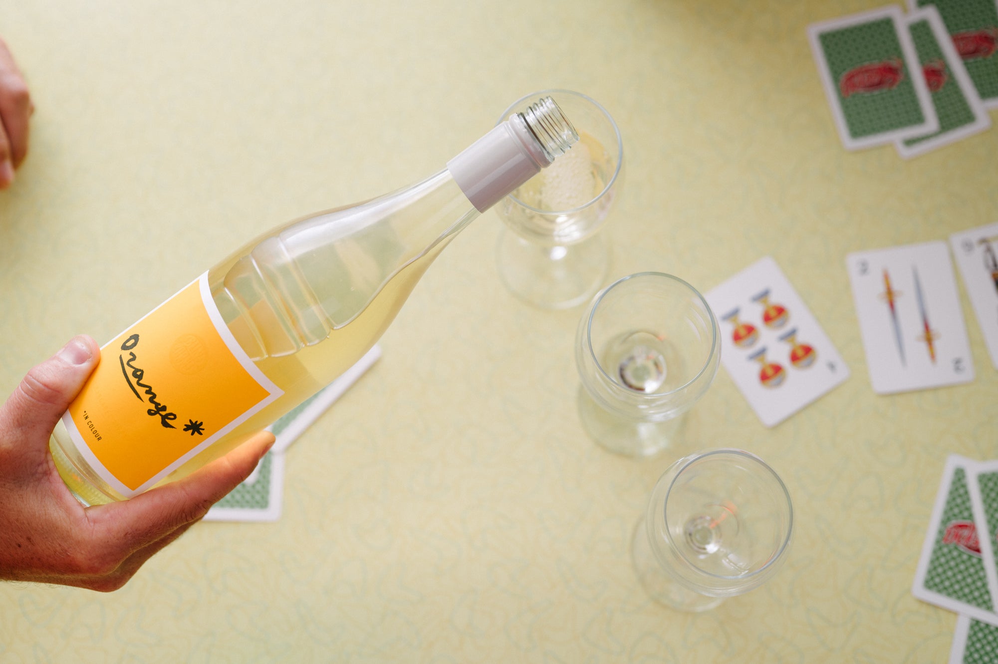 Hand holding a bottle of white wine with two glasses on a table with playing cards.